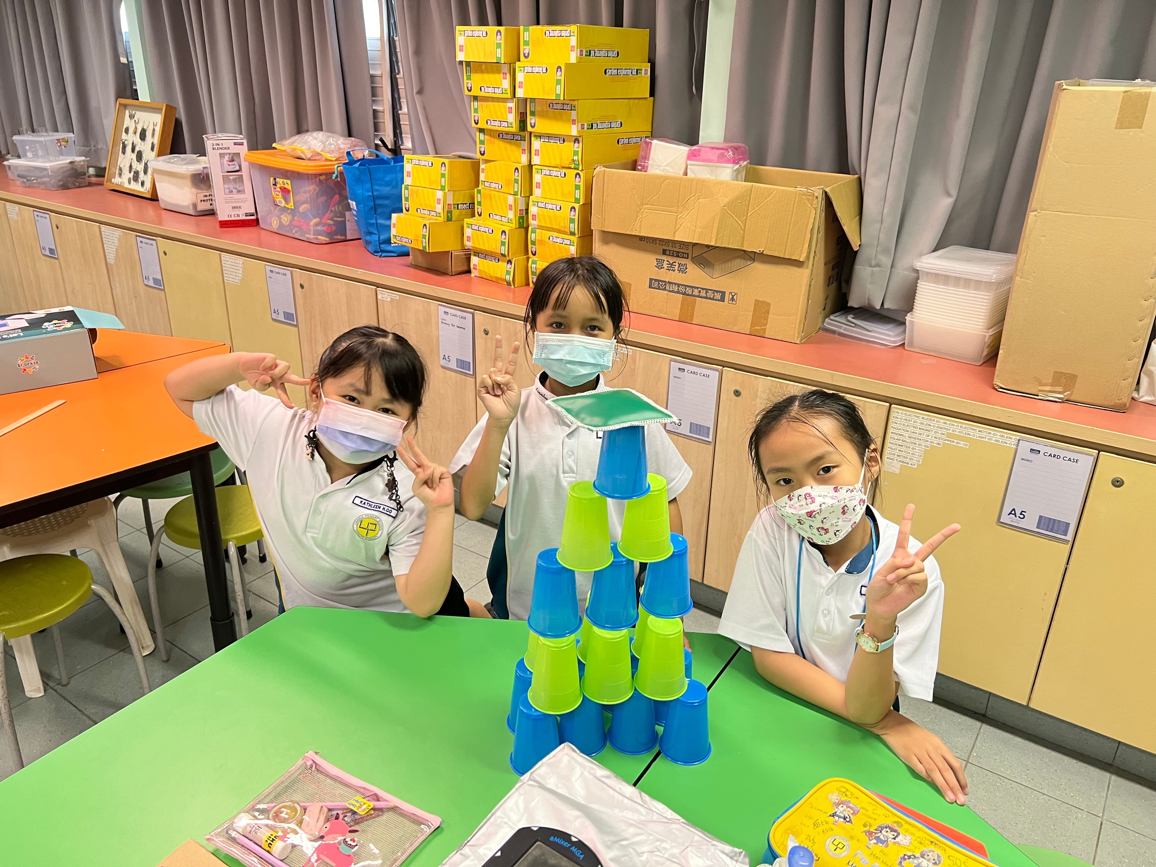 Three masked schoolgirls pose with peace signs beside a pyramid of stacked blue and green plastic cups on a green table.